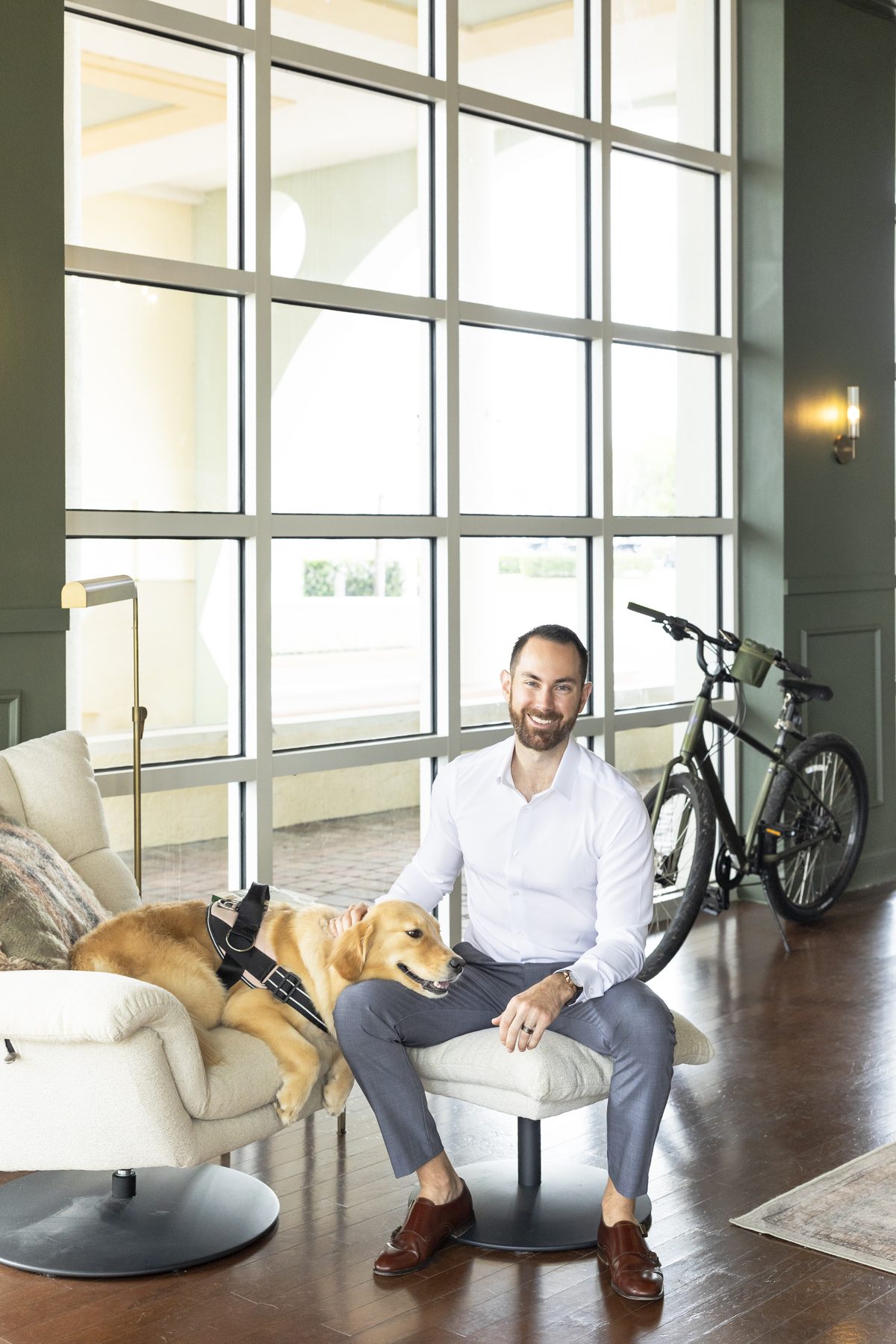 Jesse Plunkett standing by the tall office windows with his golden retriever, Nala, on the chair beside him